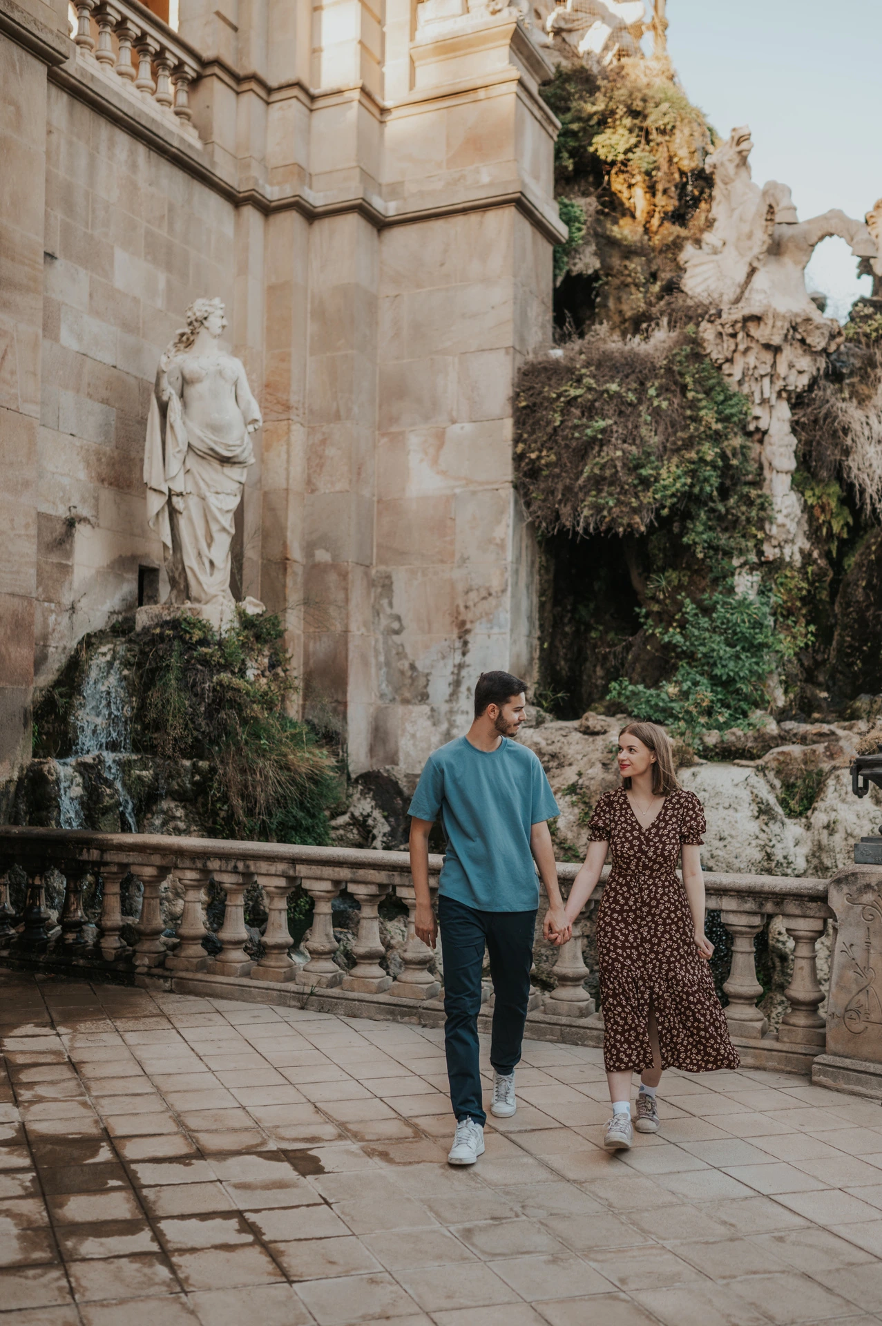 The couple walking together on the stone paving beside the fountain and waterfall during their couple photoshoot in Barcelona.