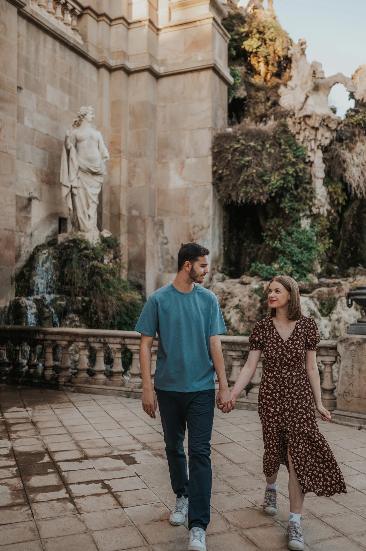 The couple walking together on the stone paving beside the fountain and waterfall during their couple photoshoot in Barcelona.