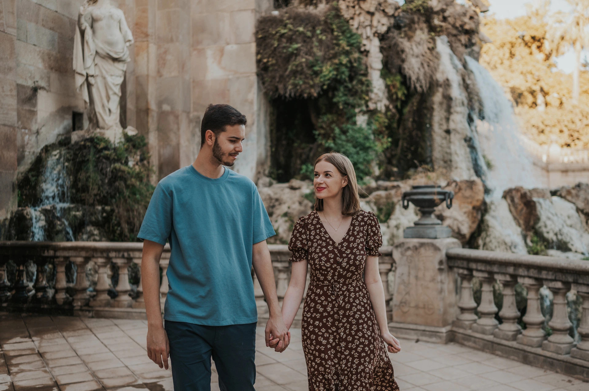 A walk past the waterfall | Barcelona couple photoshoot The couple walking together on the stone paving beside the fountain and waterfall during their couple photoshoot in Barcelona.