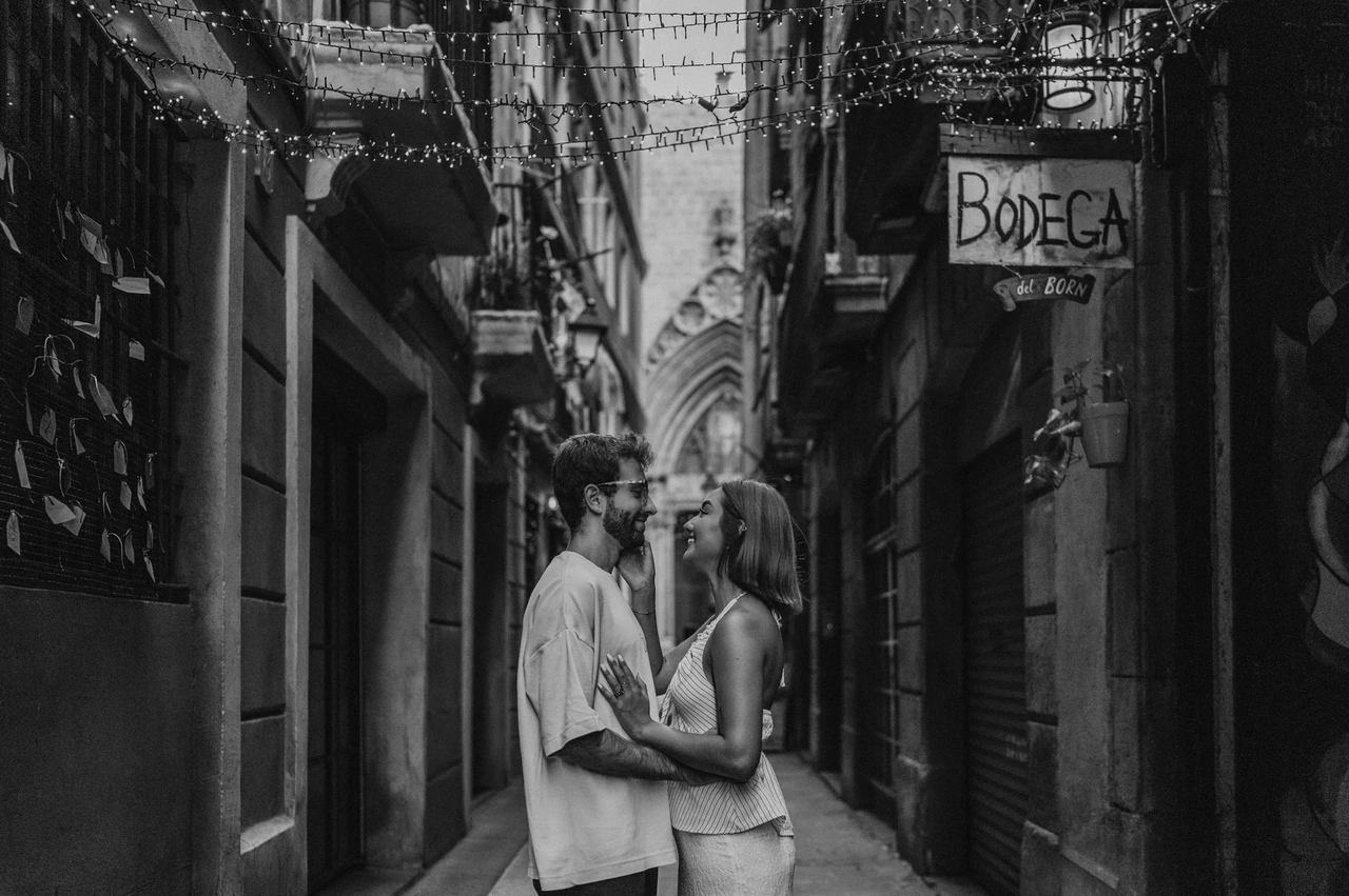 An embrace in the Bodega del Born lane in black and white | Barcelona couple photoshoot Black-and-white photograph of the couple embracing in a narrow lane beneath hanging decorations and a Bodega del Born sign during their couple photoshoot in Barcelona.