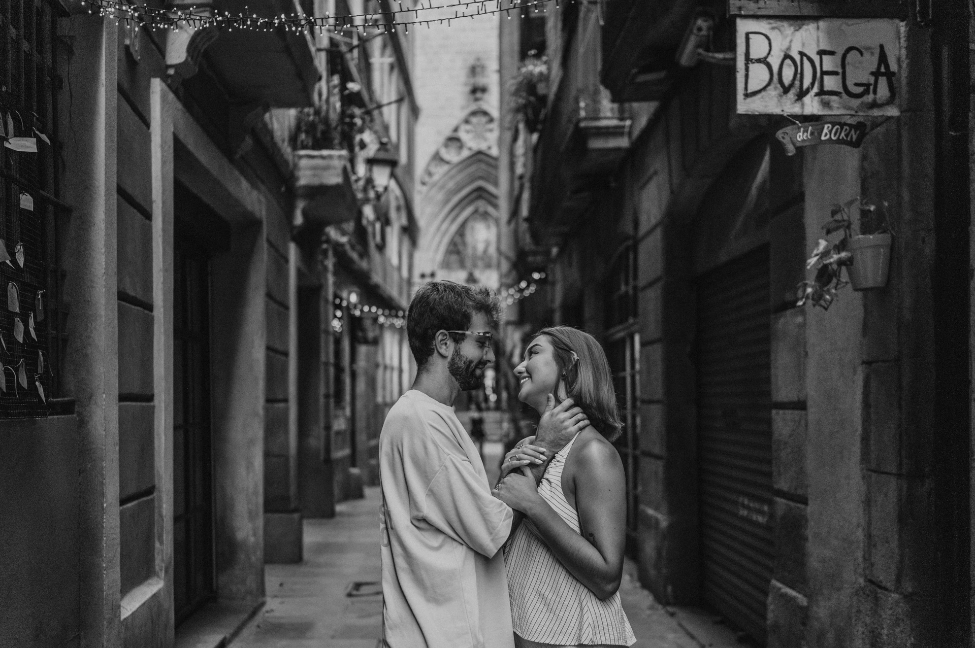 Black-and-white photograph of the couple embracing in a narrow lane below a Bodega del Born sign and hanging decorations during their couple photoshoot in Barcelona.