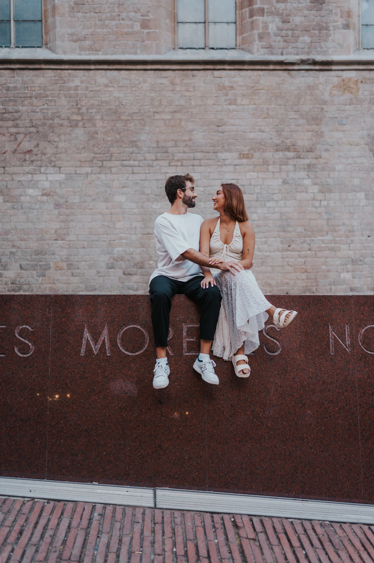 The couple sitting together on the top edge of a red metal wall against pale brickwork during their couple photoshoot in Barcelona.