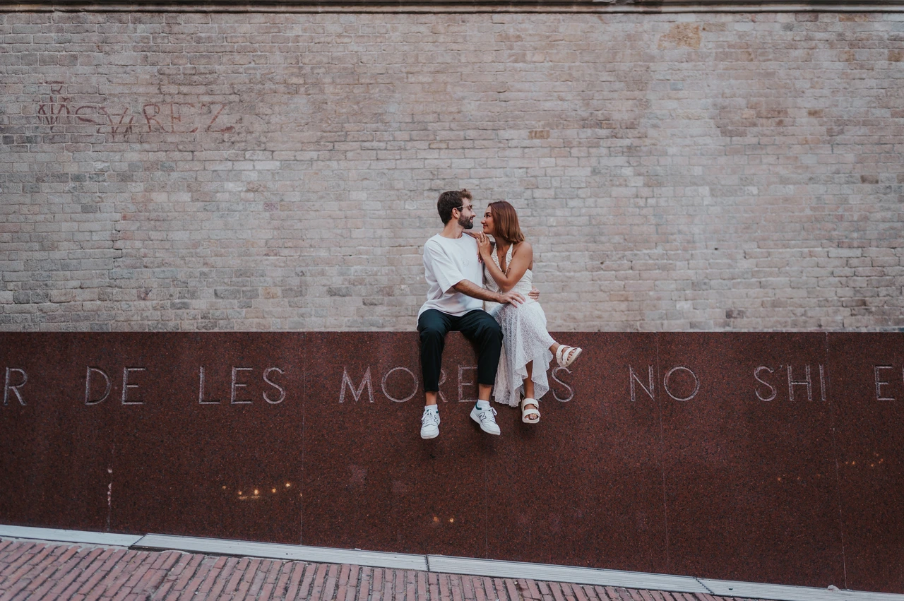 A quiet portrait against the brick wall | Barcelona couple photoshoot A wide view of the couple sitting close together on a red metal wall beneath pale brickwork during their couple photoshoot in Barcelona.