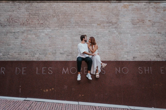 A wide view of the couple sitting close together on a red metal wall beneath pale brickwork during their couple photoshoot in Barcelona.