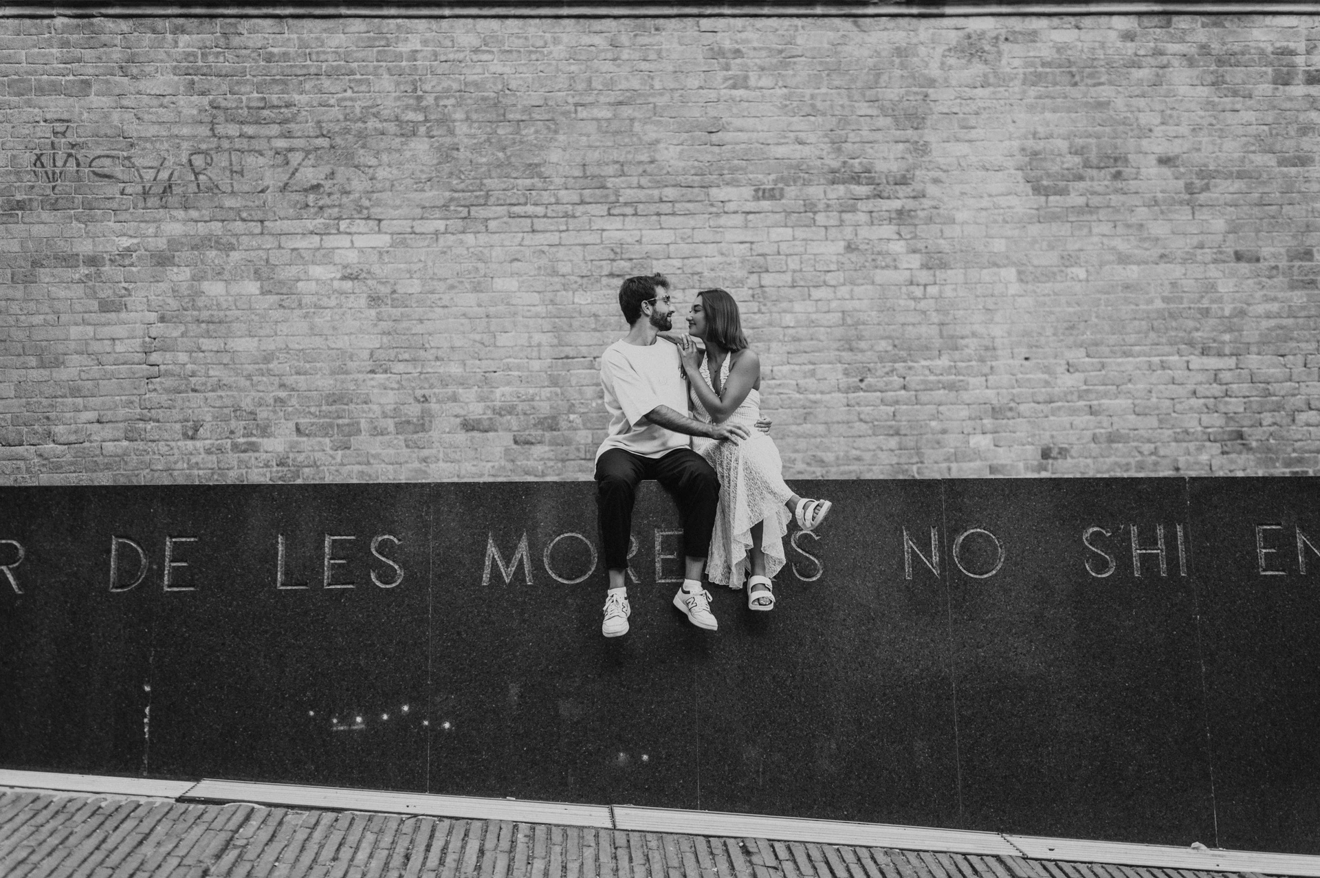 Black-and-white photograph of the couple sitting close together on a red metal wall against a broad brick façade during their couple photoshoot in Barcelona.