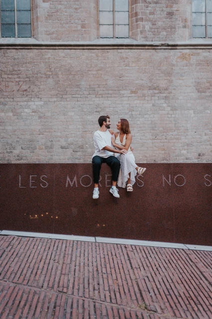 A portrait framed by brickwork and paving | Barcelona couple photoshoot The couple sitting together on a red metal wall, with the brick façade and paving around them during their couple photoshoot in Barcelona.