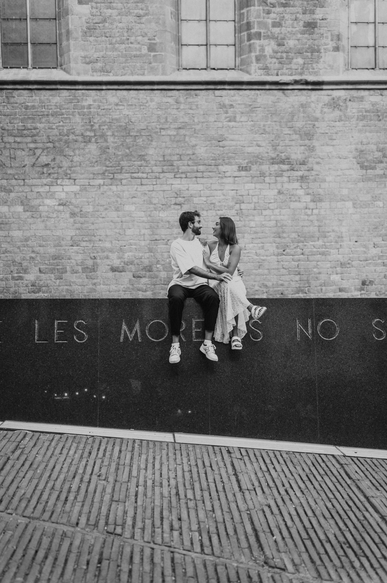 Black-and-white photograph of the couple sitting together beneath a large window on a brick façade, above the paved street during their couple photoshoot in Barcelona.