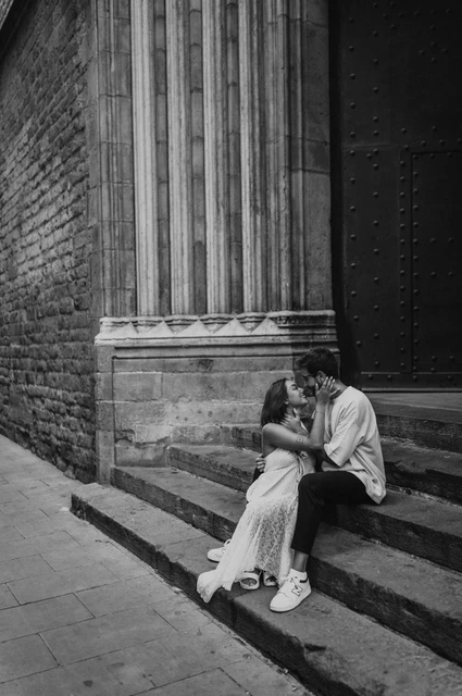 A quiet portrait on the church steps in black and white | Barcelona couple photoshoot Black-and-white photograph of the couple sitting close together on stone church steps beneath a carved stone column during their couple photoshoot in Barcelona.