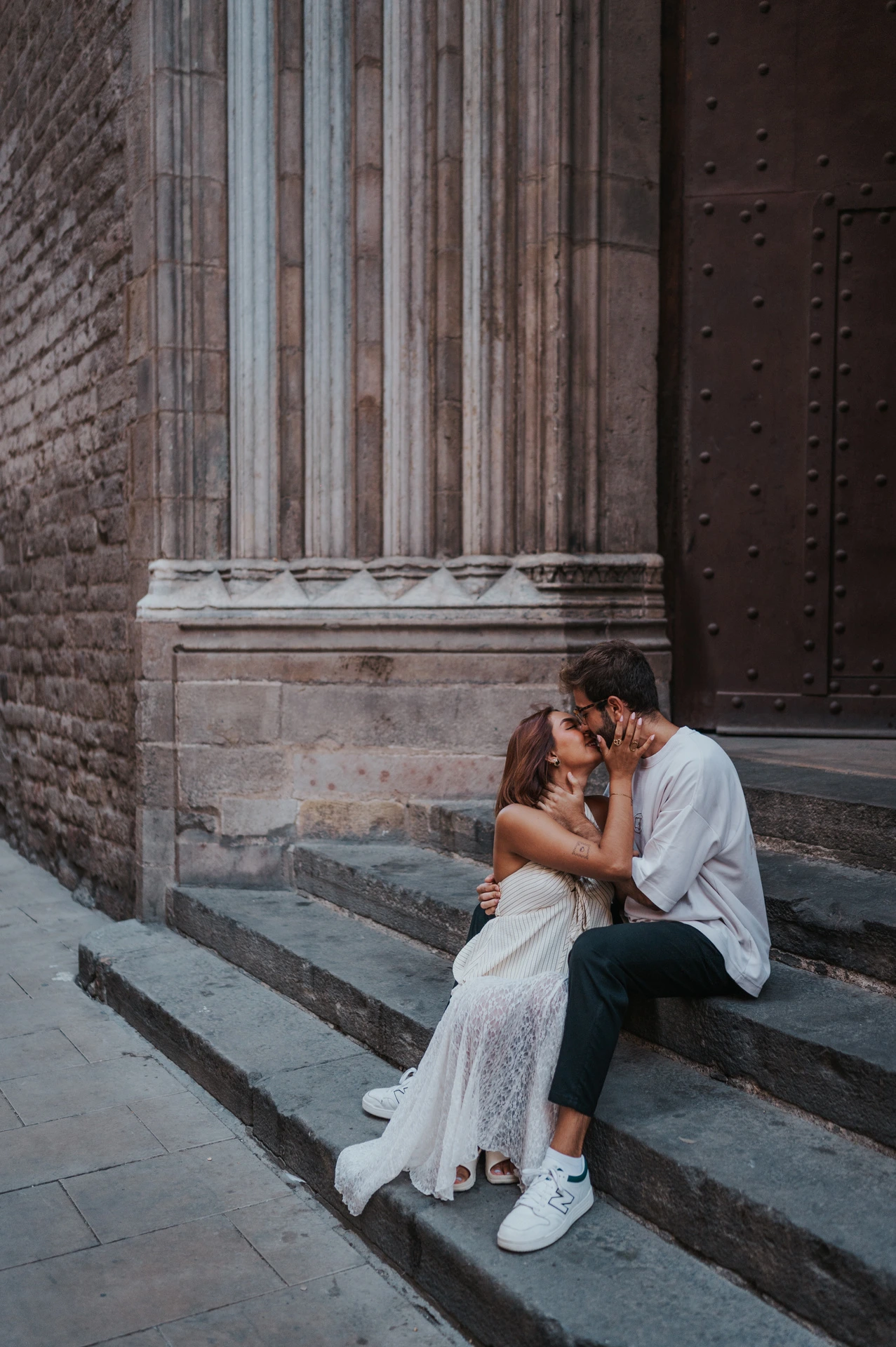 The couple kissing while sitting on stone steps beside a carved church doorway during their couple photoshoot in Barcelona.