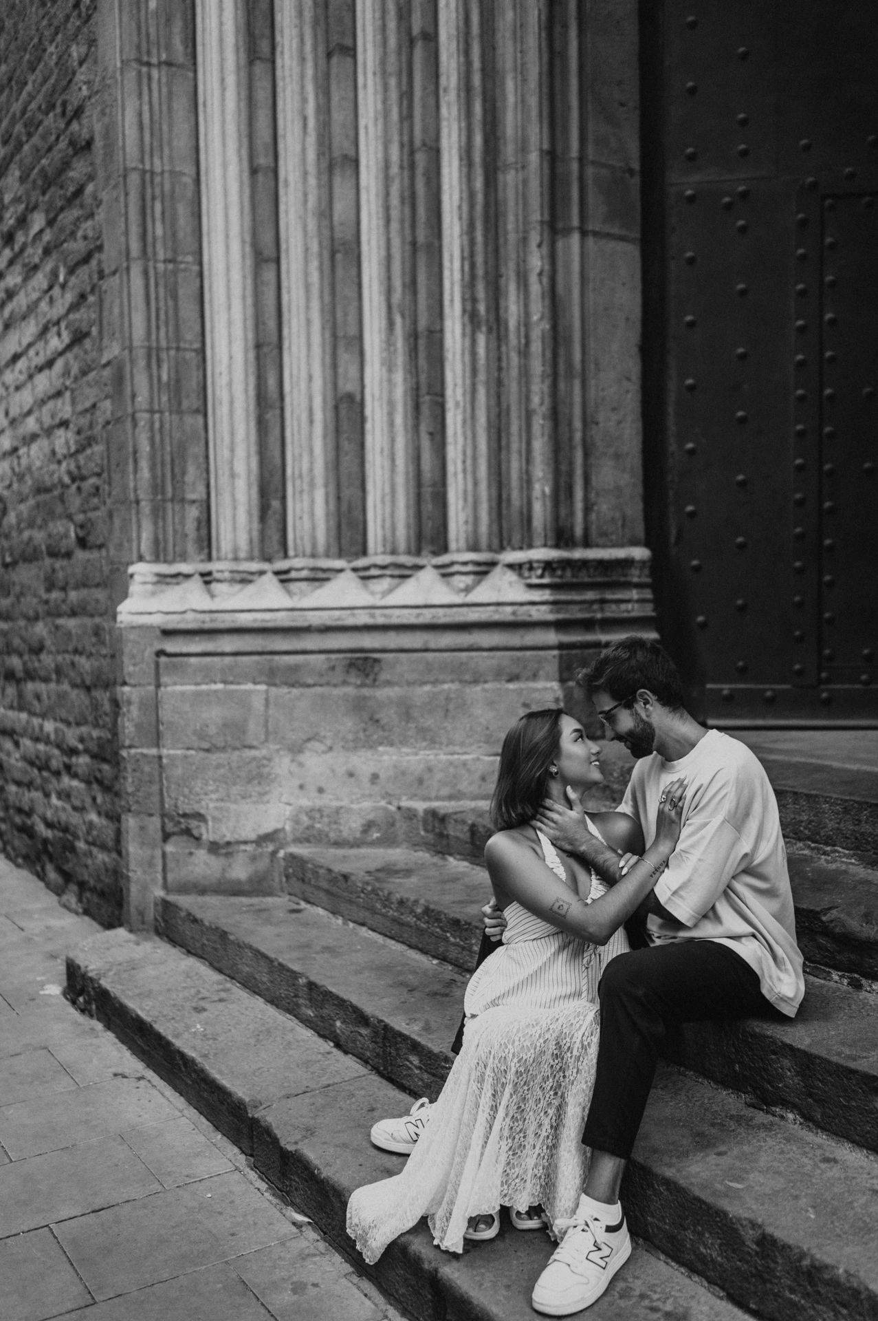 Black-and-white photograph of a close view of the couple sitting together on stone church steps beside a carved column during their couple photoshoot in Barcelona.