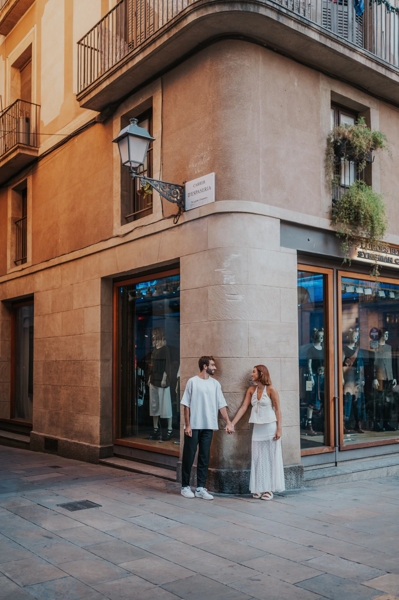 The couple holding hands beside the corner of a warm-toned building with wrought-iron balconies during their couple photoshoot in Barcelona.