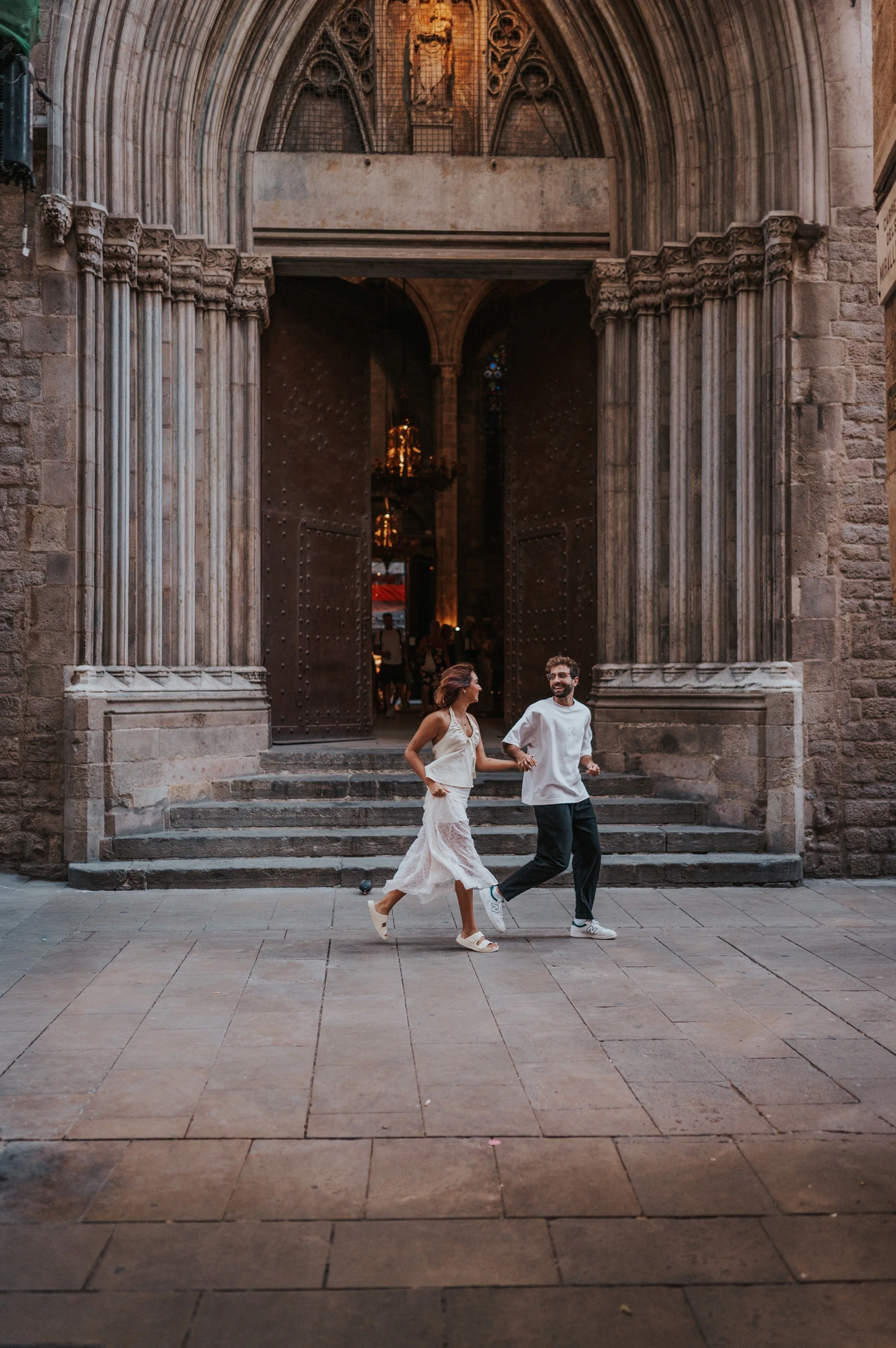 The couple moving across a paved square in front of a tall pointed church doorway during their couple photoshoot in Barcelona.