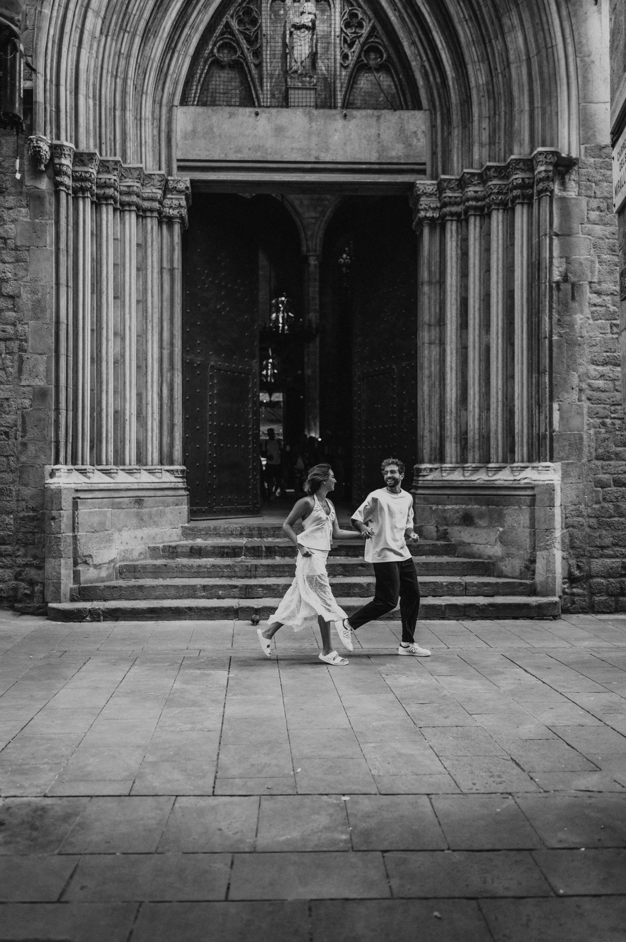 Black-and-white photograph of the couple holding hands in front of the open wooden doors and carved arch of a Gothic church during their couple photoshoot in Barcelona.