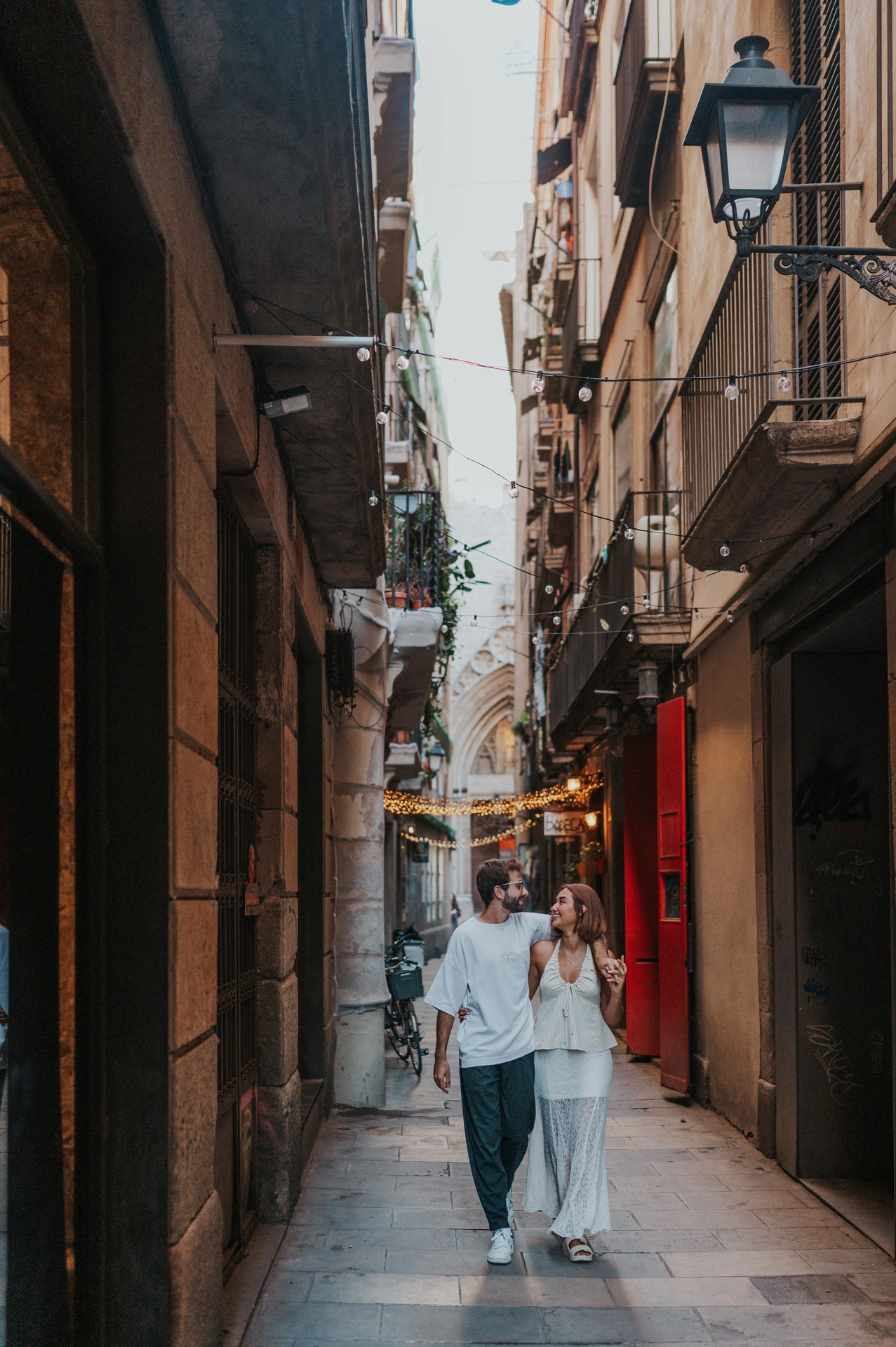 The couple walking side by side through a narrow lane beneath old lamps and small balconies during their couple photoshoot in Barcelona.
