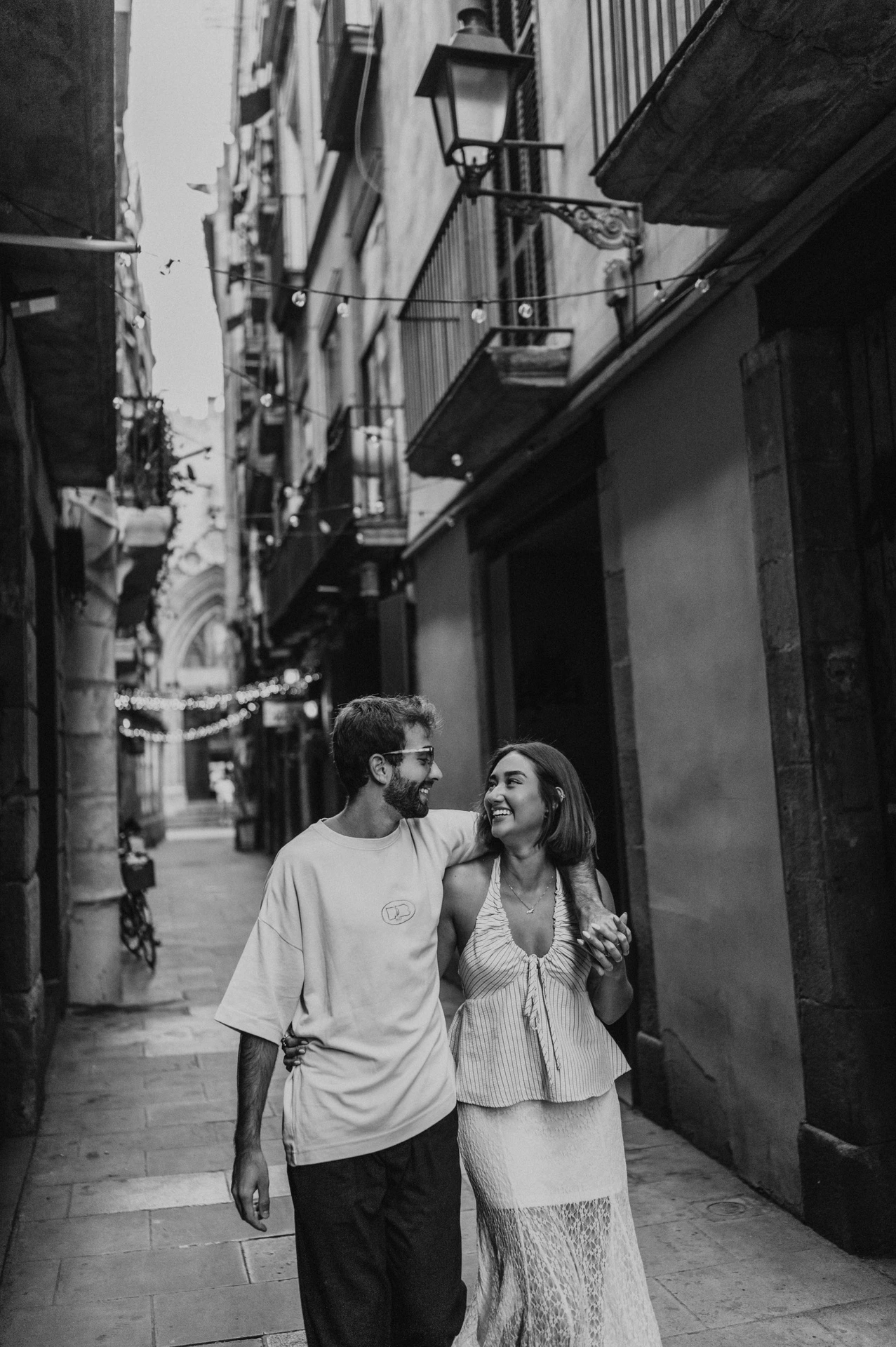 Black-and-white photograph of the couple smiling at each other while walking together beneath a street lamp in a narrow lane during their couple photoshoot in Barcelona.