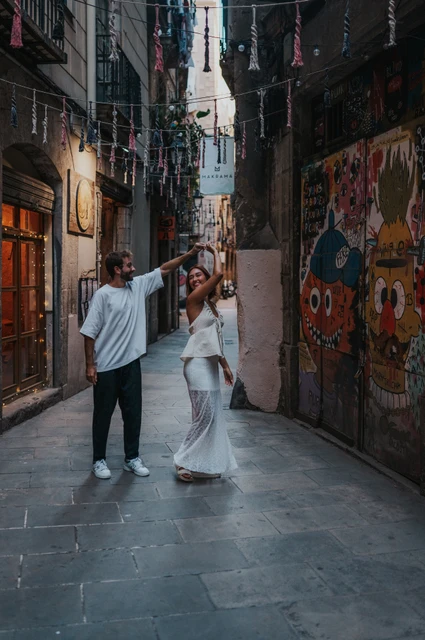 The man lifting their joined hands as his partner turns in a narrow lane beside a colourful wall mural during their couple photoshoot in Barcelona.