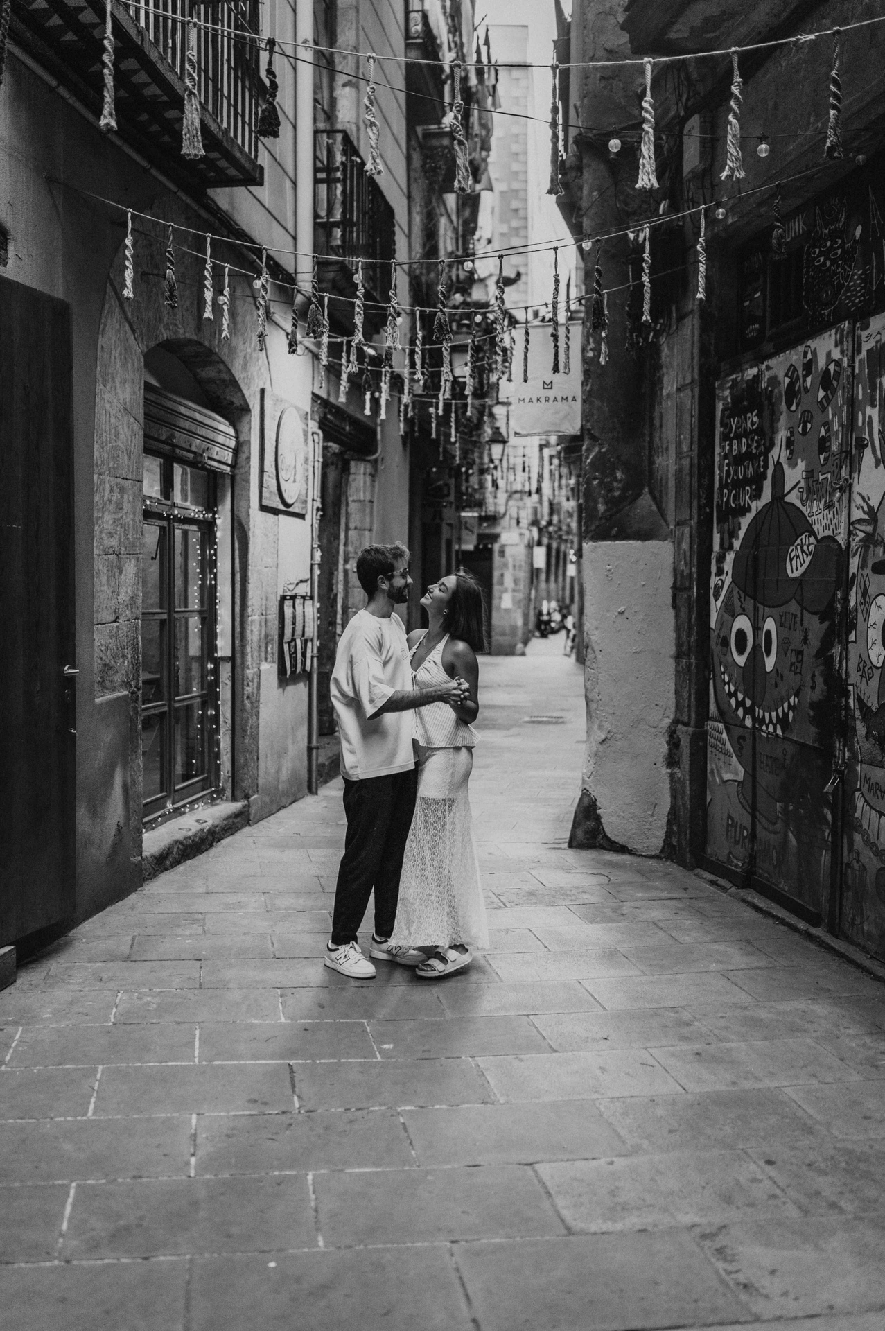 Black-and-white photograph of the couple embracing in a narrow lane beneath hanging decorations beside a wall mural during their couple photoshoot in Barcelona.