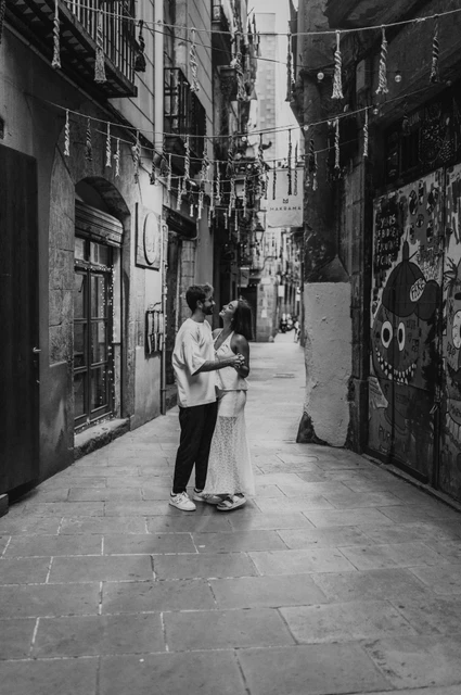 An embrace in the mural-lined lane in black and white | Barcelona couple photoshoot Black-and-white photograph of the couple embracing in a narrow lane beneath hanging decorations beside a wall mural during their couple photoshoot in Barcelona.