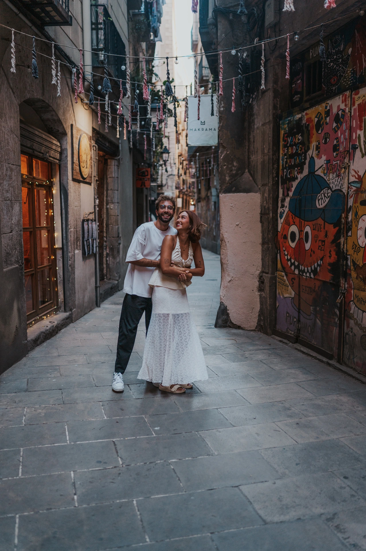 The man embracing his partner from behind in a narrow lane with a colourful mural and overhead decorations during their couple photoshoot in Barcelona.