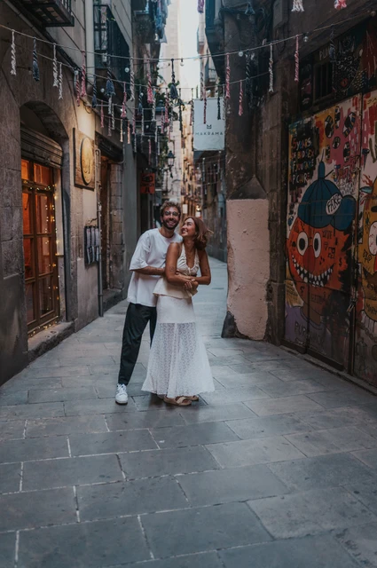 A smiling portrait in the decorated lane | Barcelona couple photoshoot The man embracing his partner from behind in a narrow lane with a colourful mural and overhead decorations during their couple photoshoot in Barcelona.