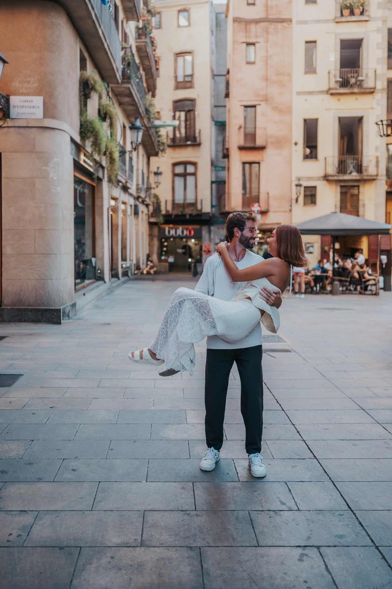 The man carrying his partner in an open paved square surrounded by warm-toned buildings during their couple photoshoot in Barcelona.