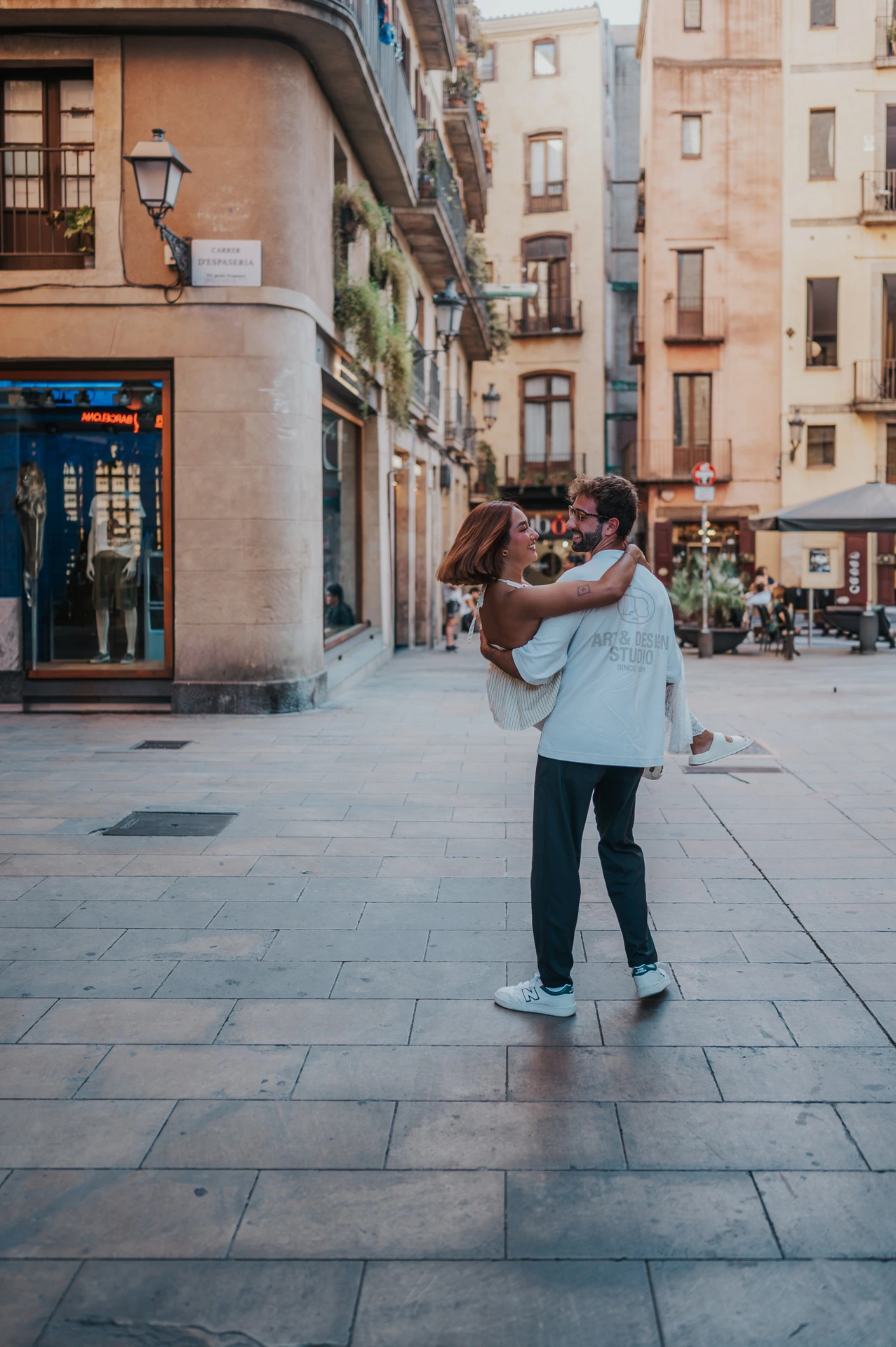 The man holding his partner in his arms in a paved square beside a corner building during their couple photoshoot in Barcelona.