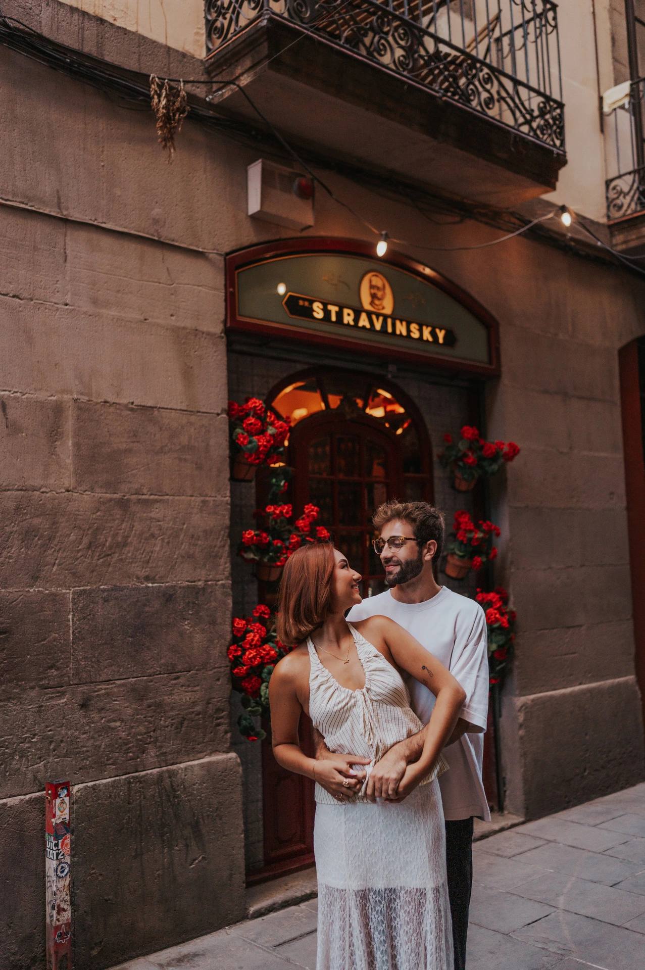 The man embracing his partner from behind beside a doorway framed with red flowers during their couple photoshoot in Barcelona.
