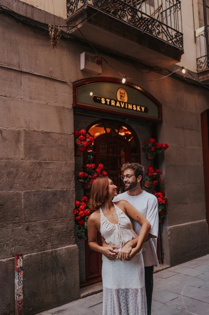 Together beside a flower-framed doorway | Barcelona couple photoshoot The man embracing his partner from behind beside a doorway framed with red flowers during their couple photoshoot in Barcelona.