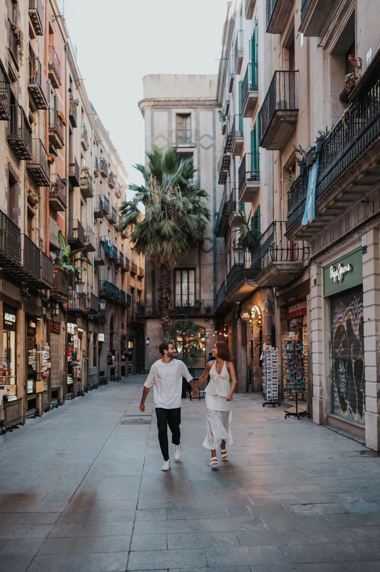 The couple walking hand in hand along a shop-lined street beneath balconies and a palm tree during their couple photoshoot in Barcelona.