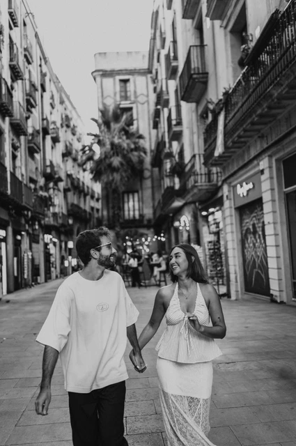 Together beneath the street’s balconies in black and white | Barcelona couple photoshoot Black-and-white photograph of the couple walking close together beneath balconies on an old town shopping street during their couple photoshoot in Barcelona.
