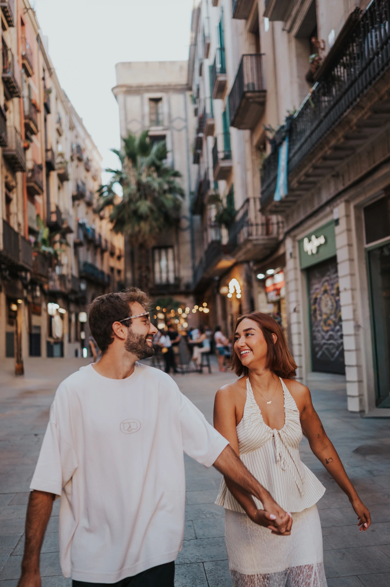 A close view of the couple smiling at each other while walking along a shop-lined street during their couple photoshoot in Barcelona.
