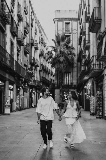 A stroll between the old shopfronts in black and white | Barcelona couple photoshoot Black-and-white photograph of the couple walking toward the camera together on a street lined with balconies and shops during their couple photoshoot in Barcelona.