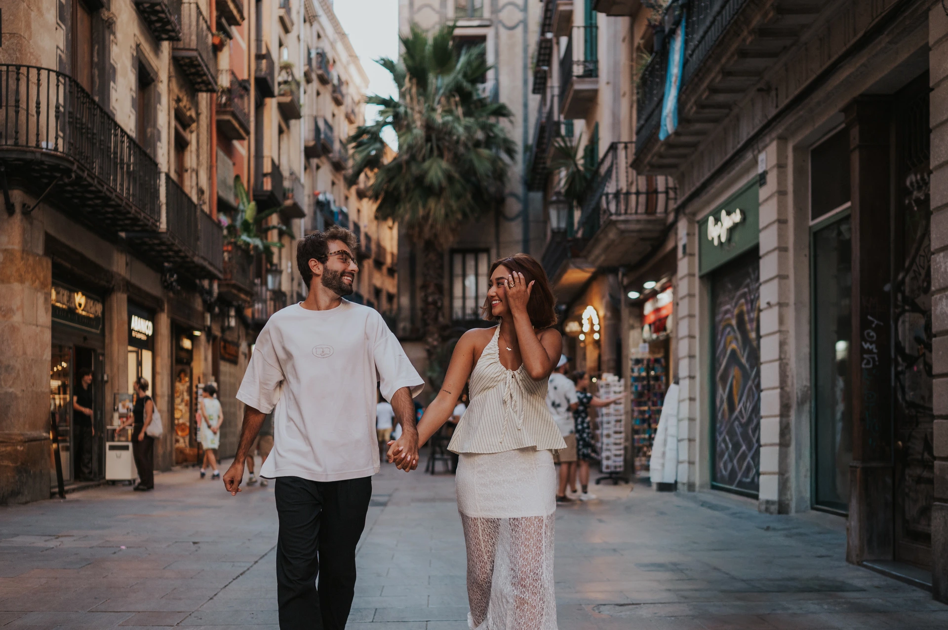 A wide street portrait together | Barcelona couple photoshoot The couple walking hand in hand and looking at each other on a shop-lined street during their couple photoshoot in Barcelona.