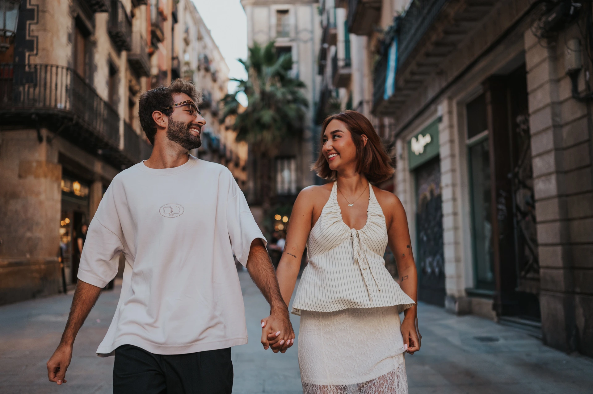 The couple smiling while holding hands on a street with shops and balconies behind them during their couple photoshoot in Barcelona.