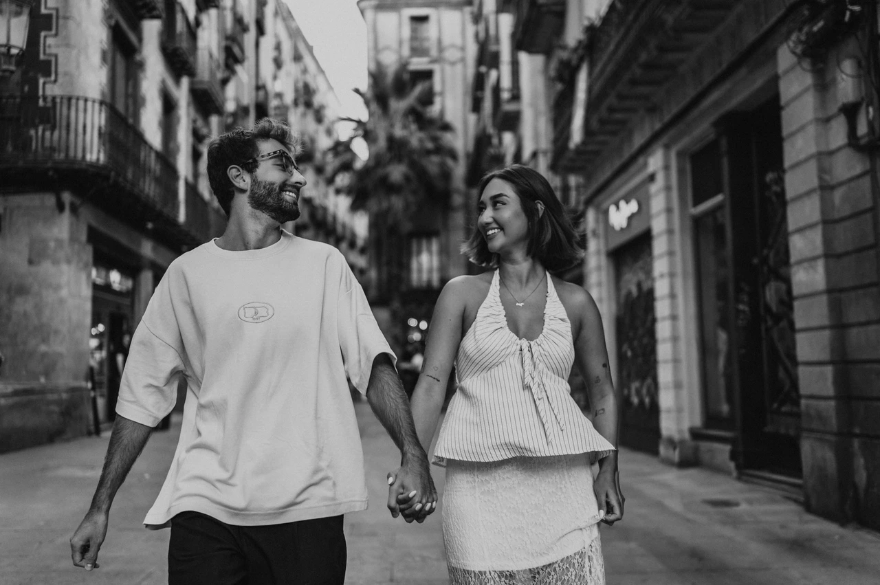 A close street portrait together in black and white | Barcelona couple photoshoot Black-and-white photograph of the couple looking at each other while walking hand in hand along a shop-lined street during their couple photoshoot in Barcelona.