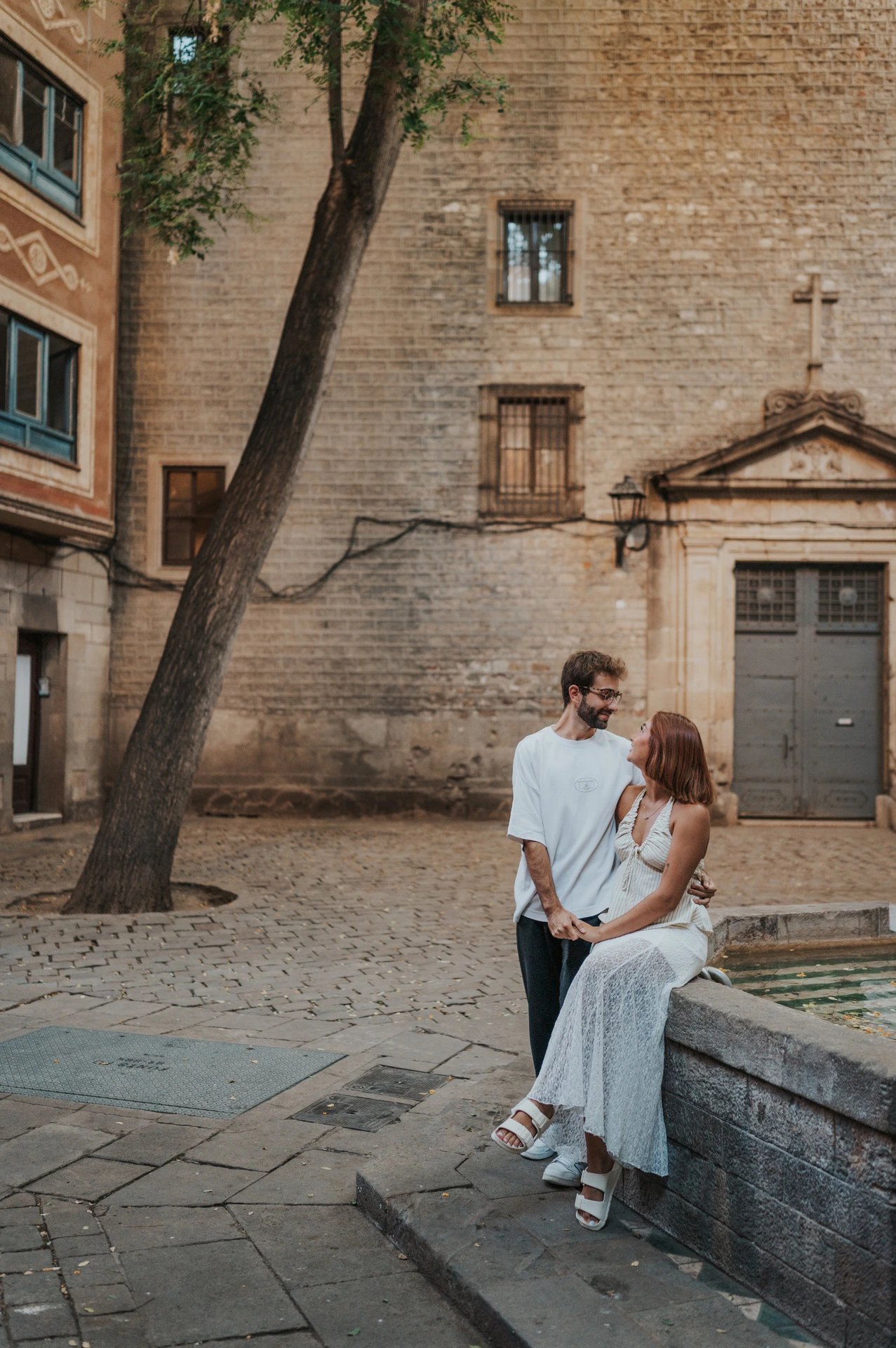 The couple beside the stone edge of a fountain in a paved square, with a tree and historic façades behind them during their couple photoshoot in Barcelona.