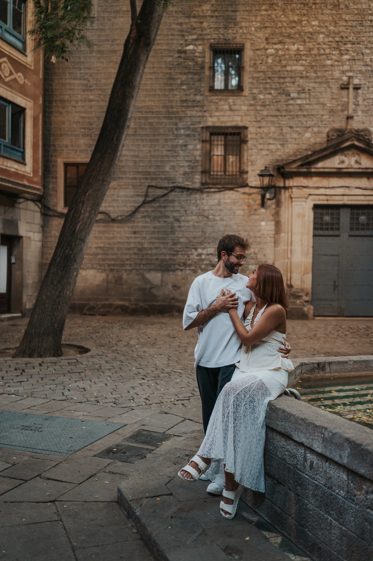 The woman sitting on the stone edge of a fountain while her partner stands close beside her in a historic square during their couple photoshoot in Barcelona.