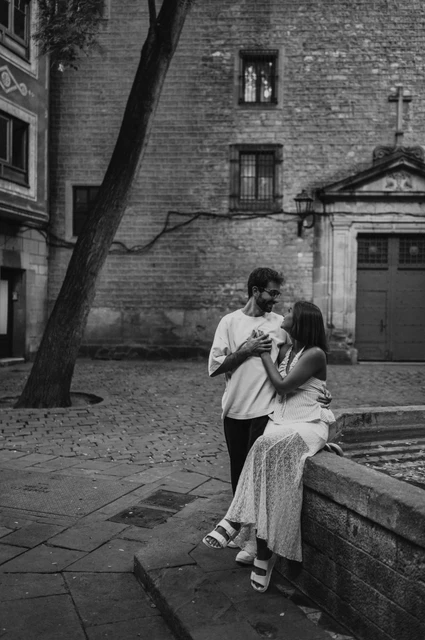 A quiet portrait beside the fountain in black and white | Barcelona couple photoshoot Black-and-white photograph of the couple standing and sitting close together beside the stone edge of a fountain in a paved square during their couple photoshoot in Barcelona.