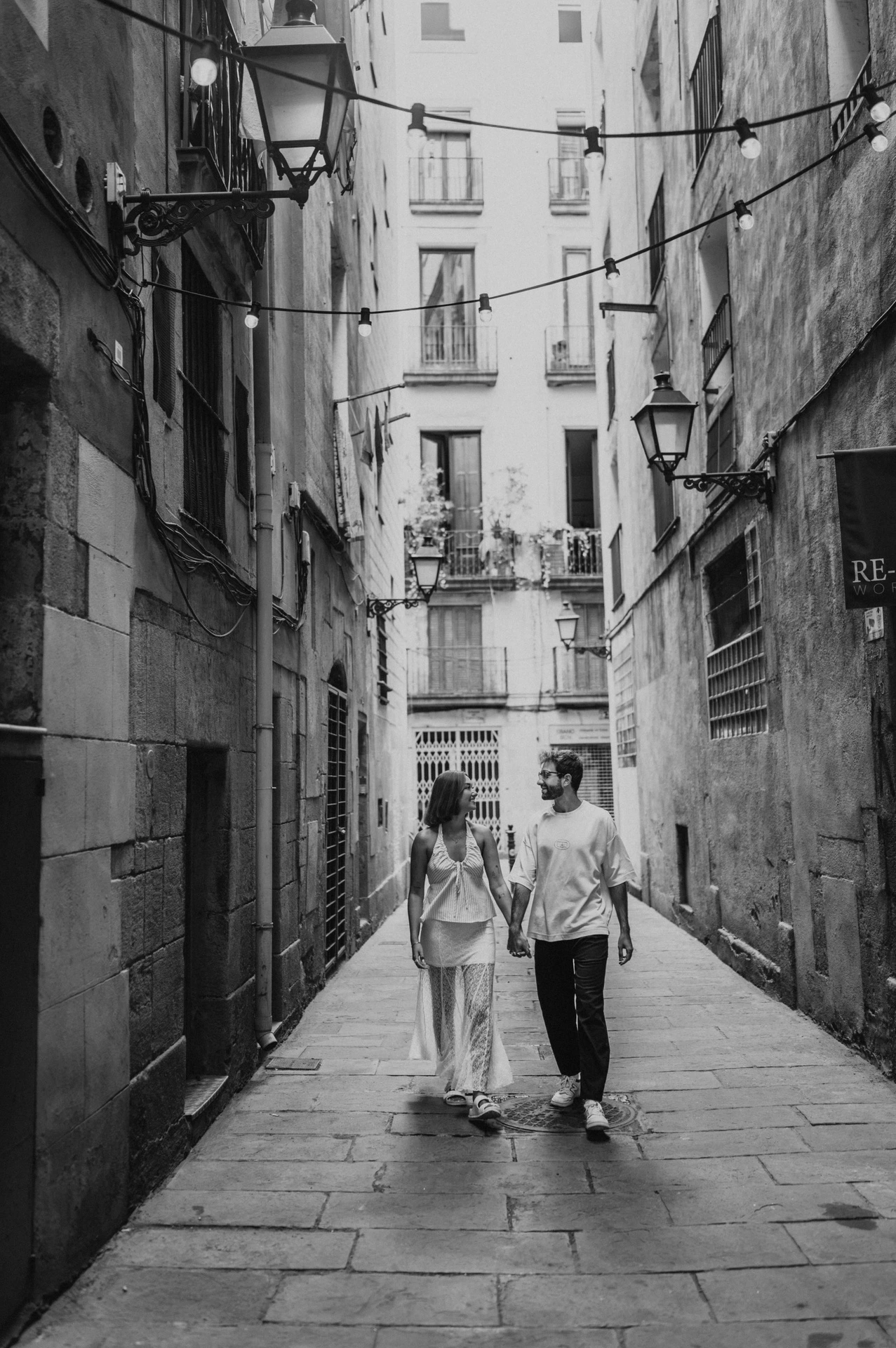 Black-and-white photograph of the couple walking together along a narrow lane between historic stone buildings during their couple photoshoot in Barcelona.