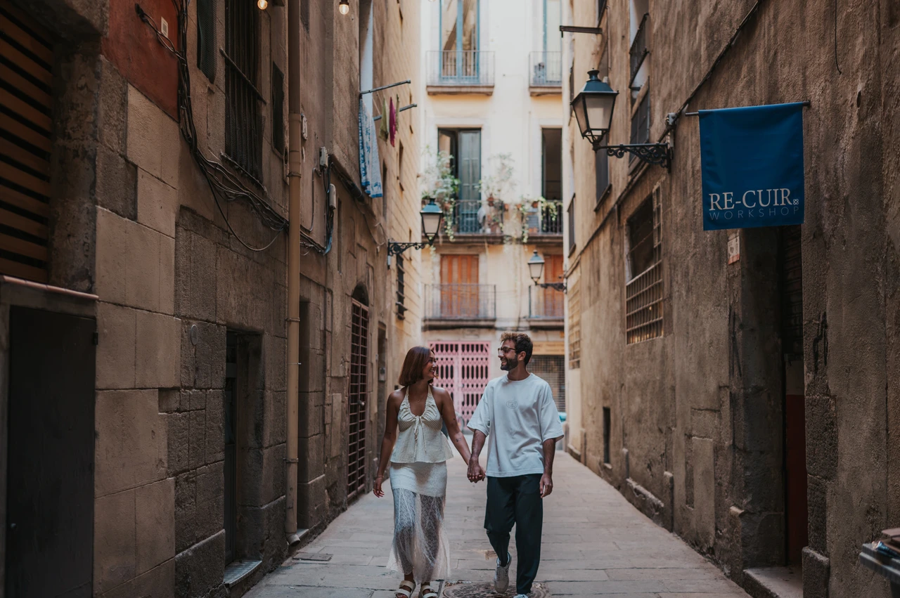 A wide view of the old town lane | Barcelona couple photoshoot A wide view of the couple holding hands in a narrow lane, with stone façades and old lamps around them during their couple photoshoot in Barcelona.