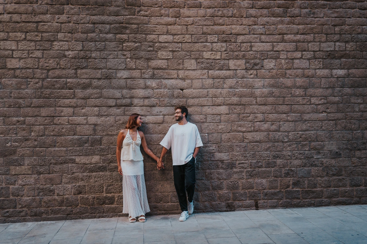 A portrait against textured stone | Barcelona couple photoshoot A wide view of the couple holding hands against a broad wall of textured stone blocks during their couple photoshoot in Barcelona.