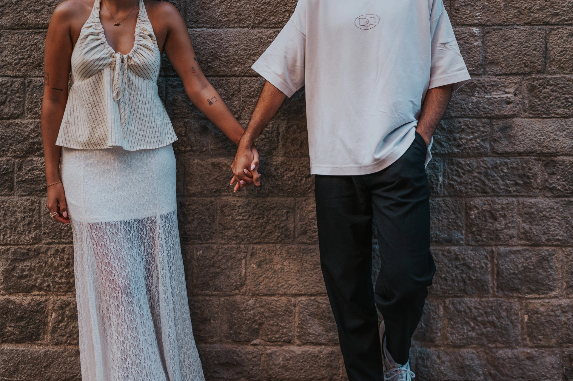 A close view of the couple’s joined hands and light outfits against a textured stone wall during their couple photoshoot in Barcelona.