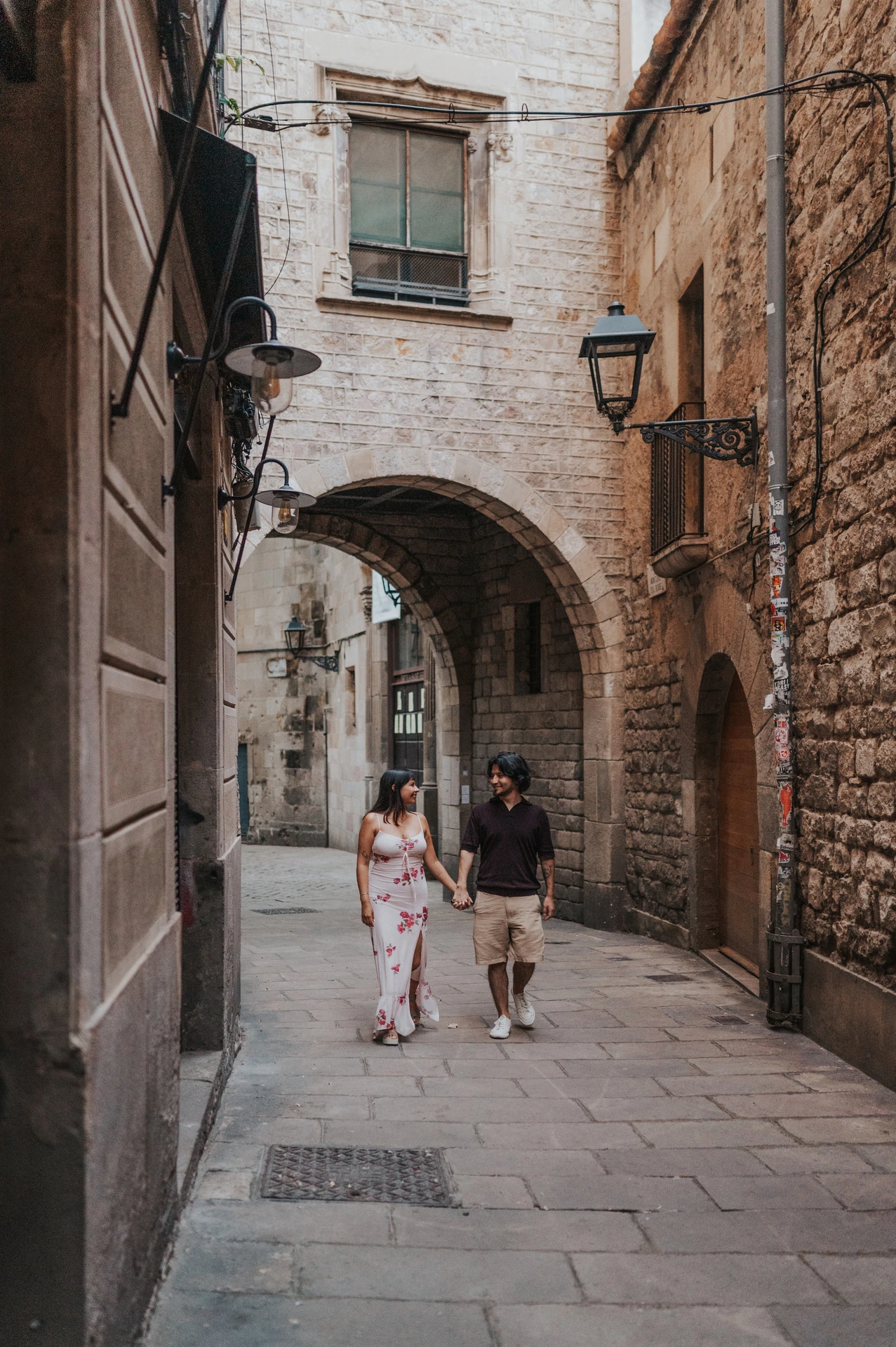 The couple walking hand in hand along a narrow stone lane beneath an arch in the Gothic Quarter during their couple photoshoot in Barcelona.