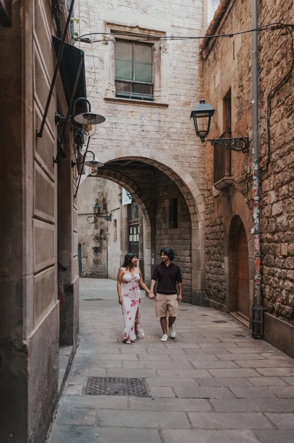 A walk along an arched Gothic Quarter lane | Barcelona couple photoshoot The couple walking hand in hand along a narrow stone lane beneath an arch in the Gothic Quarter during their couple photoshoot in Barcelona.