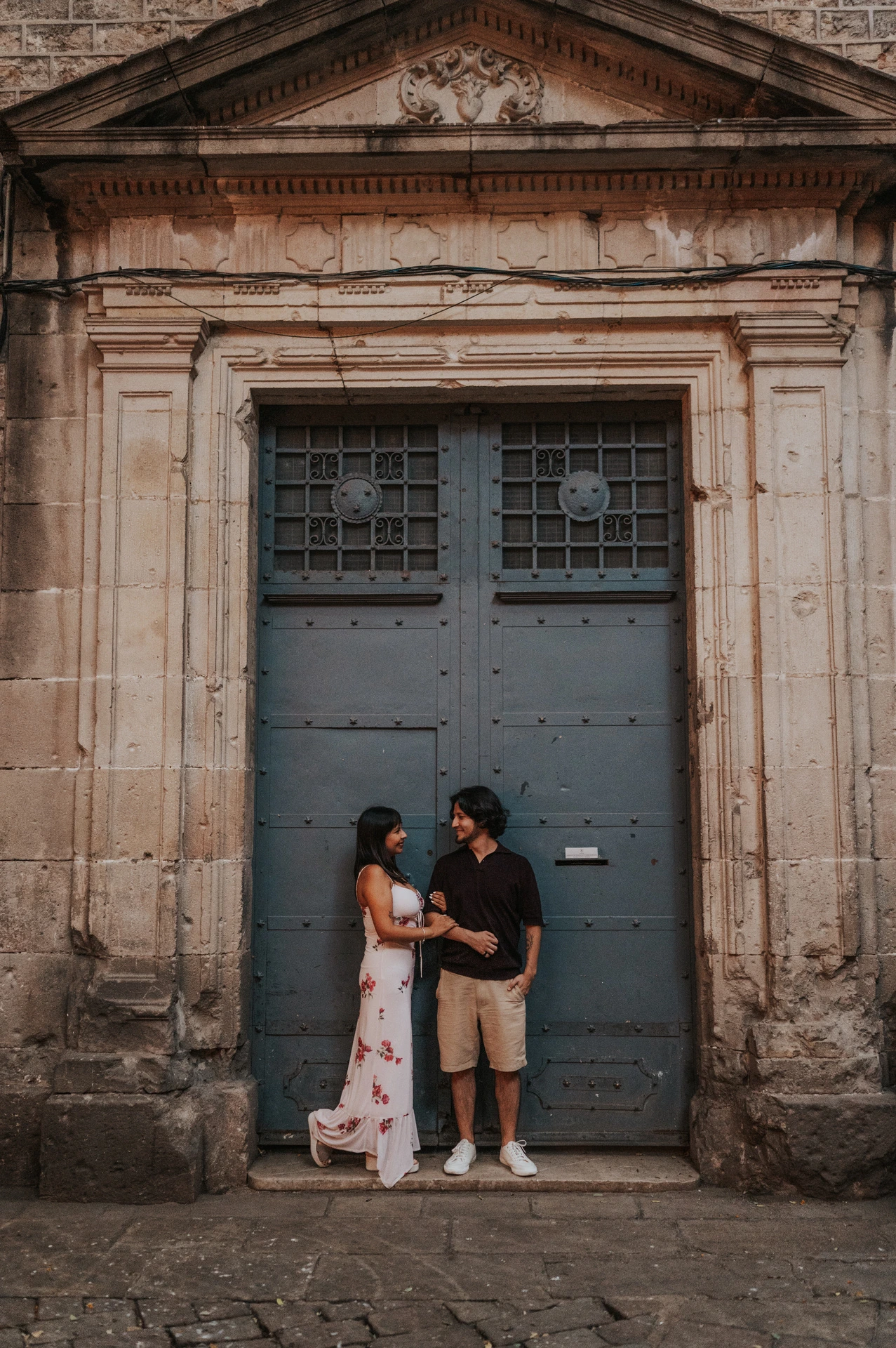 The couple standing close together and looking at each other beneath the stone pediment of an old blue-grey doorway in the Gothic Quarter during their couple photoshoot in Barcelona.