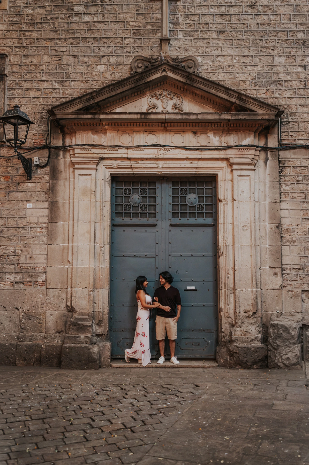 A wide portrait of the couple beneath a cross and carved stone pediment around an old blue-grey doorway in the Gothic Quarter during their couple photoshoot in Barcelona.
