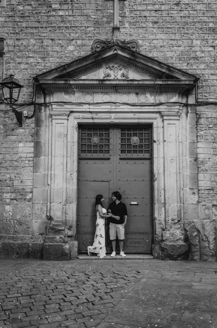 Together beside an old blue-grey doorway in black and white | Barcelona couple photoshoot Black-and-white photograph: the couple standing close together beneath a carved stone pediment around a blue-grey door in the Gothic Quarter during their couple photoshoot in Barcelona.