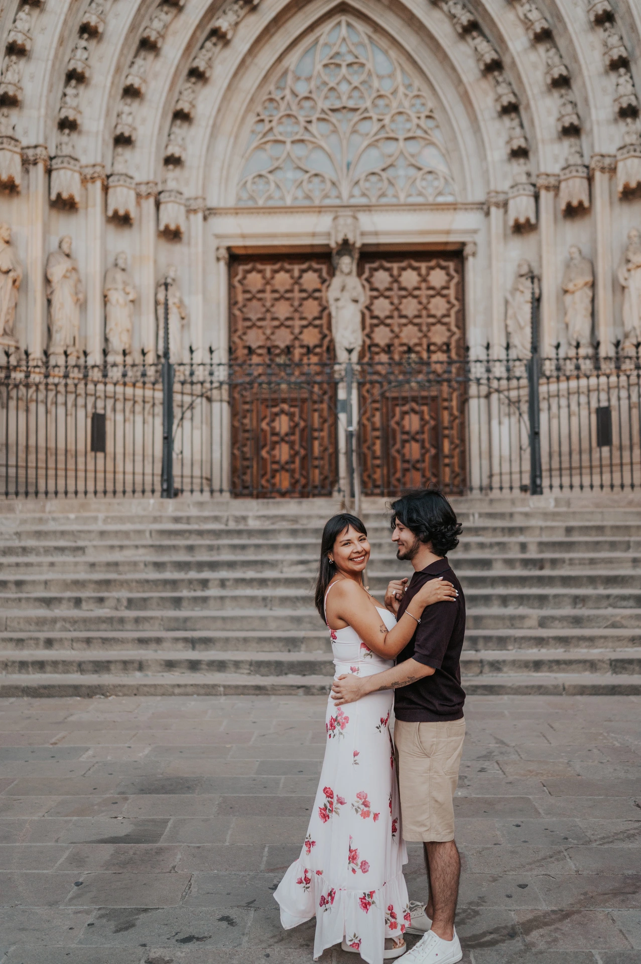 The couple embracing and smiling in front of the stone steps at Barcelona Cathedral during their couple photoshoot in Barcelona.