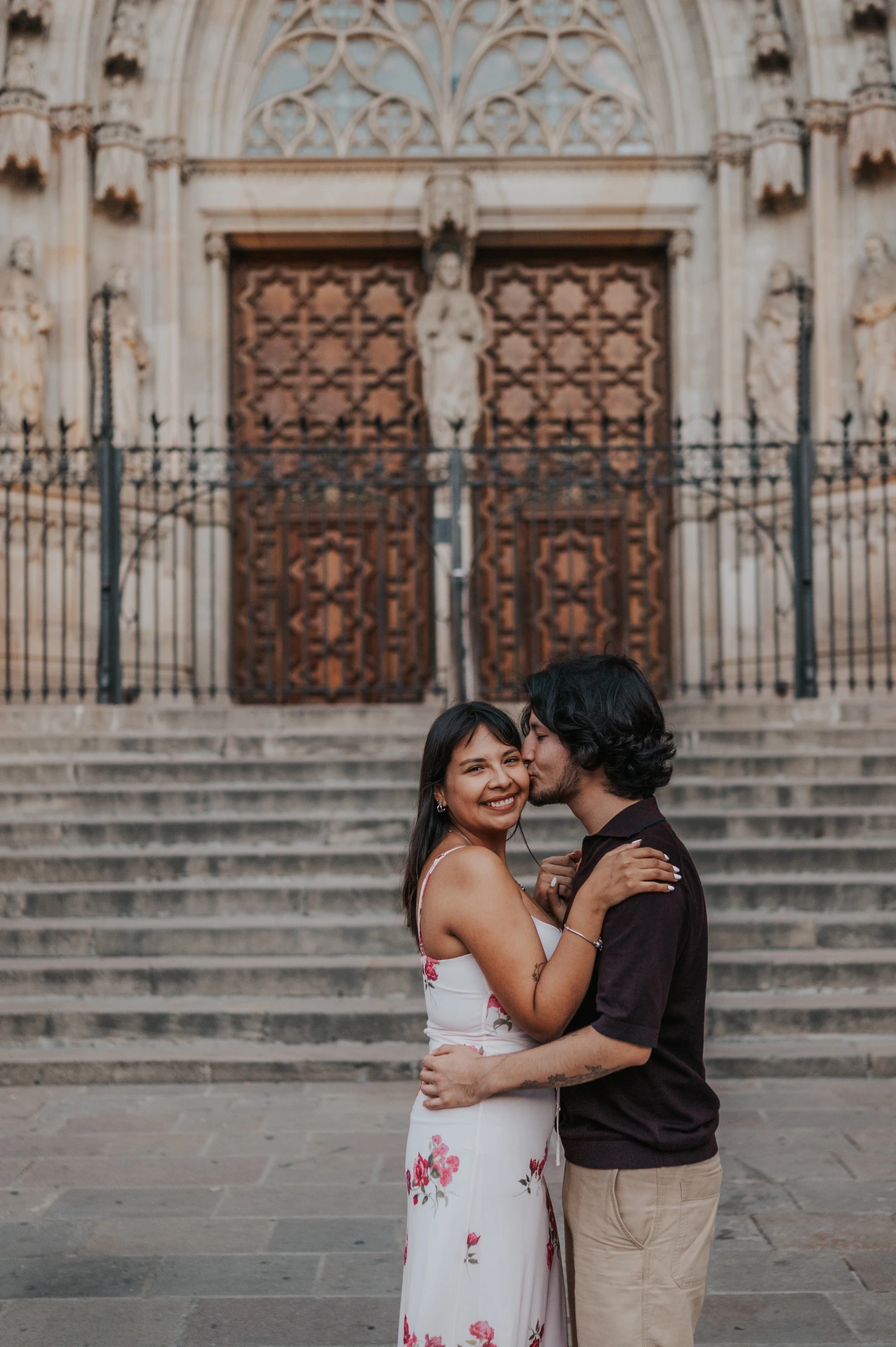 A close portrait of the man kissing his smiling partner’s cheek, with Barcelona Cathedral softly blurred behind them during their couple photoshoot in Barcelona.