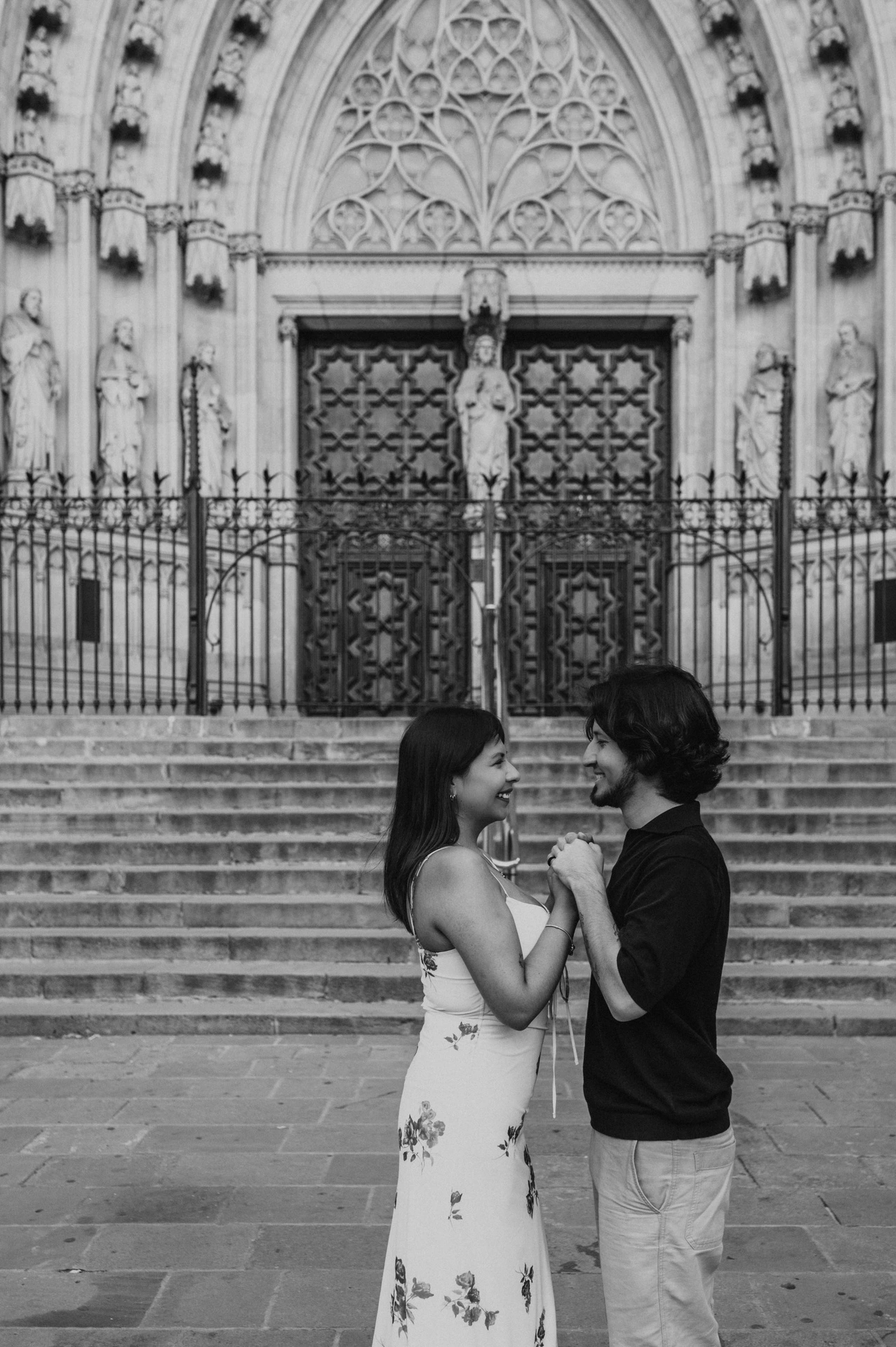 Black-and-white photograph: the couple facing each other in front of Barcelona Cathedral, with its carved doorway and railings behind them during their couple photoshoot in Barcelona.